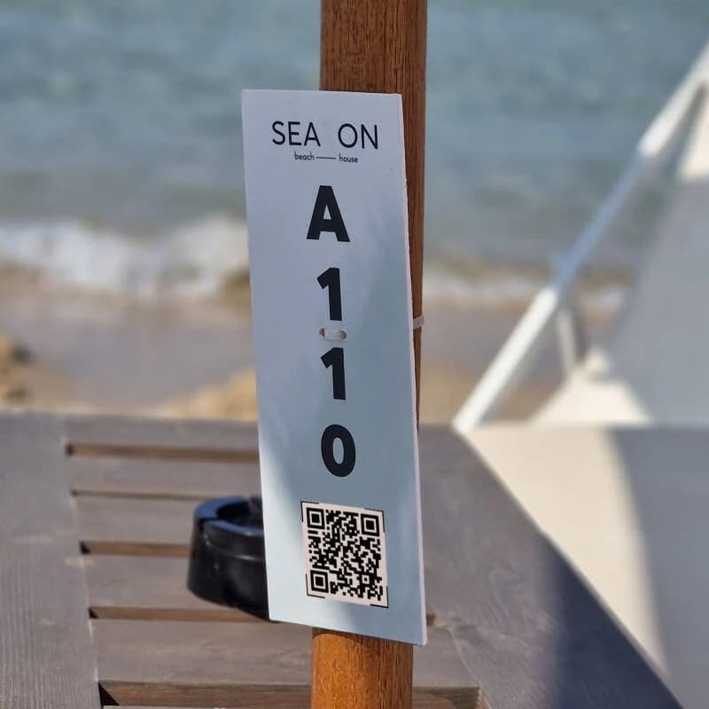 Sunbed tag with a QR code and the number A110 fastened to a beach-umbrella pole, sea in the background